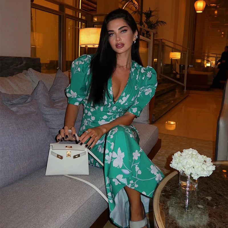 Woman in a green floral dress holding a white handbag in an indoor setting.