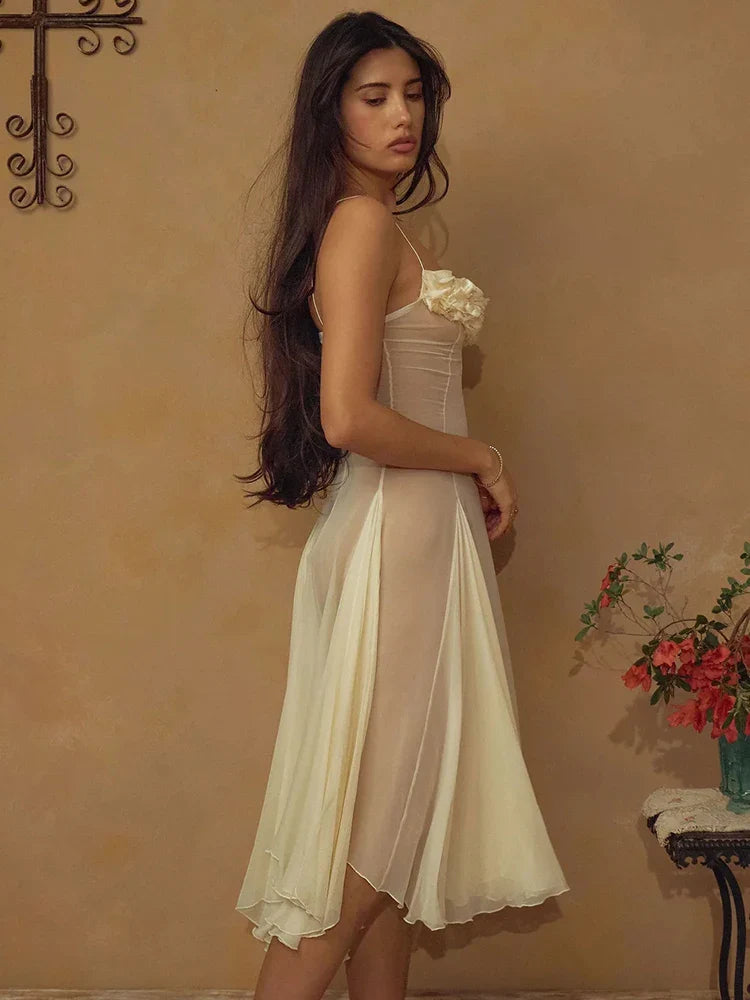 Woman in a white dress standing against a beige wall with a vase of flowers on a table.