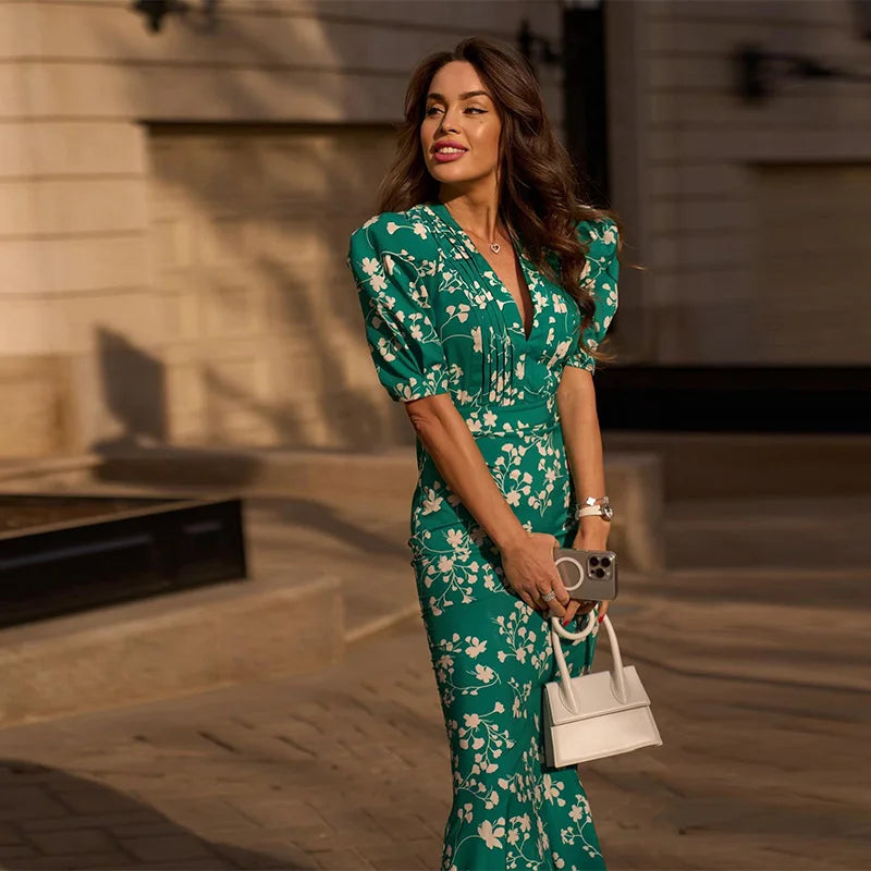 Woman in a green floral dress holding a white handbag outdoors.